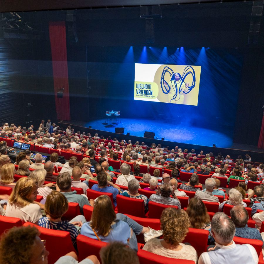 Volle theaterzaal op het podium hangt een scherm met de tekst 'Welkom Vrienden'