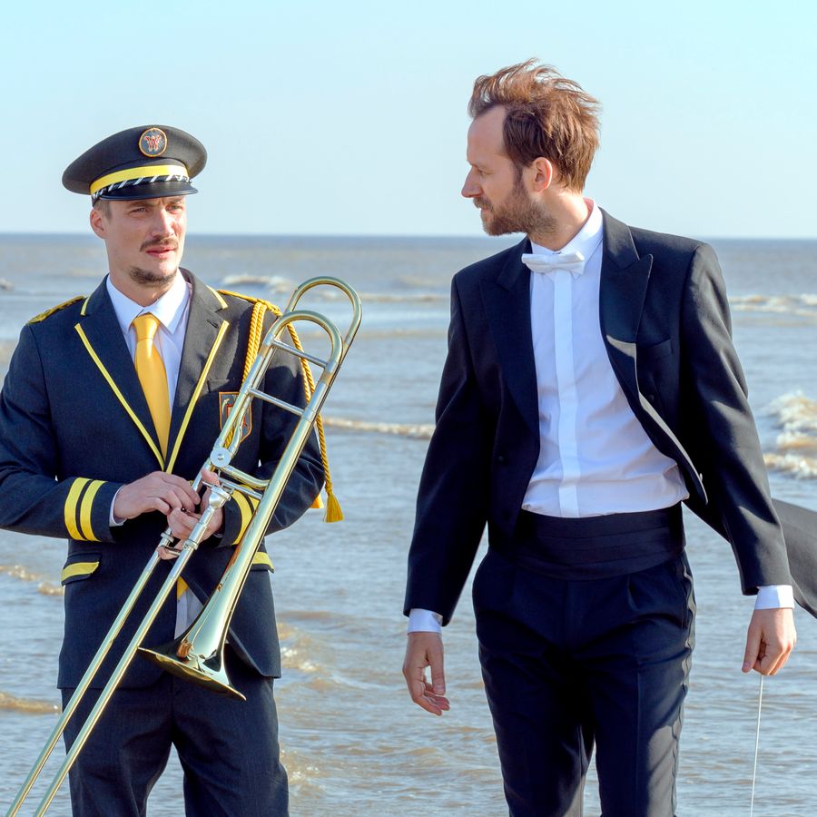 Twee mannen lopen over het strand, eentje heeft een trombone vast