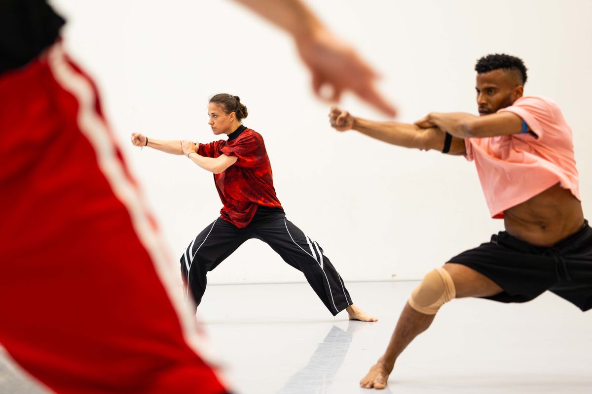 op voorgrond een been en achter twee dansers met armen gestrekt schuin naar voren