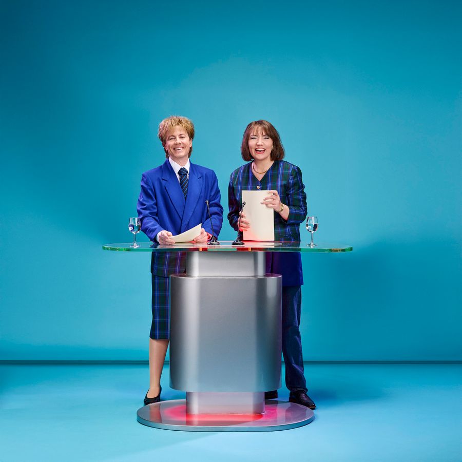 Twee Kevin van den Berg en Manon Blaauw  als nieuwslezers bij spreekstoelpersonen achter een nieuwsdesk