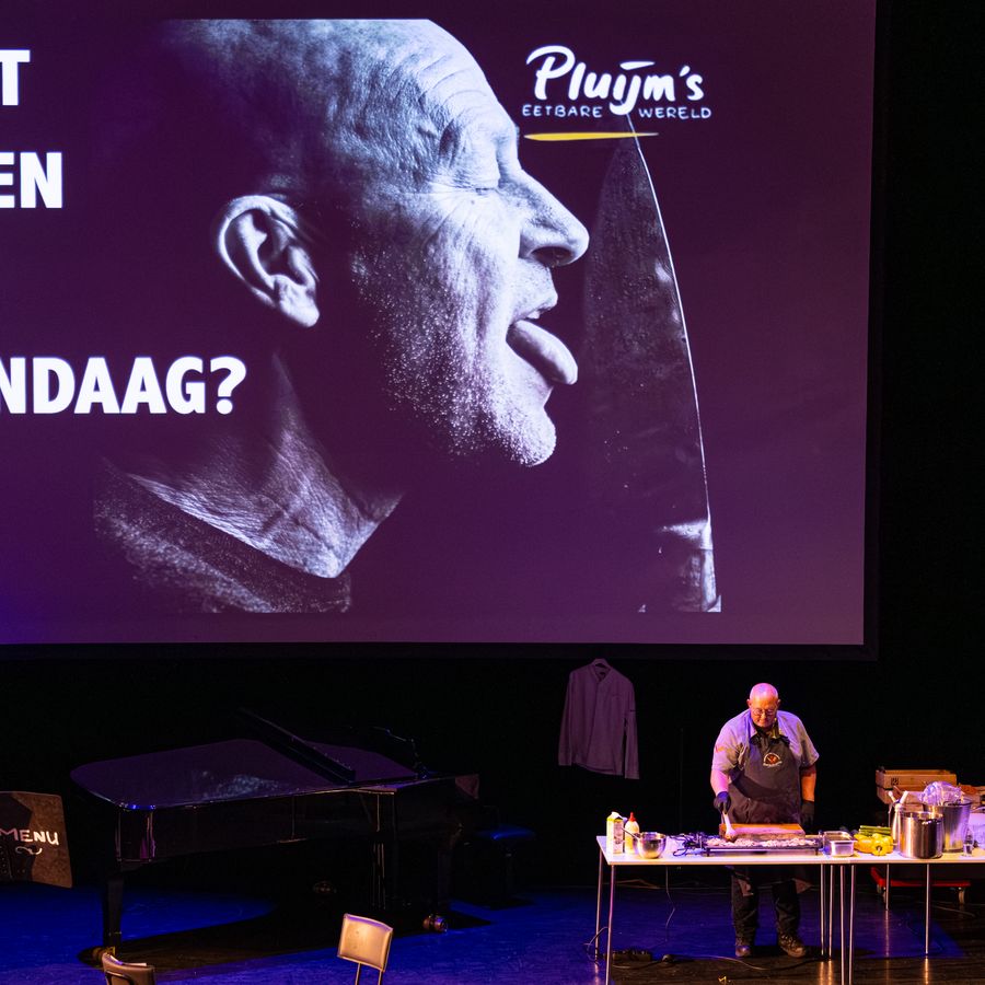 Rene Pluijm op het podium achter een tafel, op de achtergrond een scherm met de tekst WAT ETEN WE VANDAAG?
