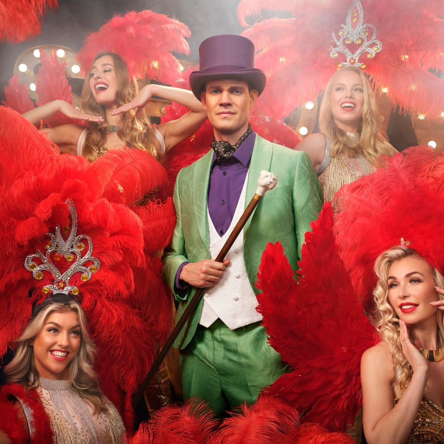 Tim Fransen in groen pak met paarse hoed. Omringd door showgirls met rode veren op het hoofd.