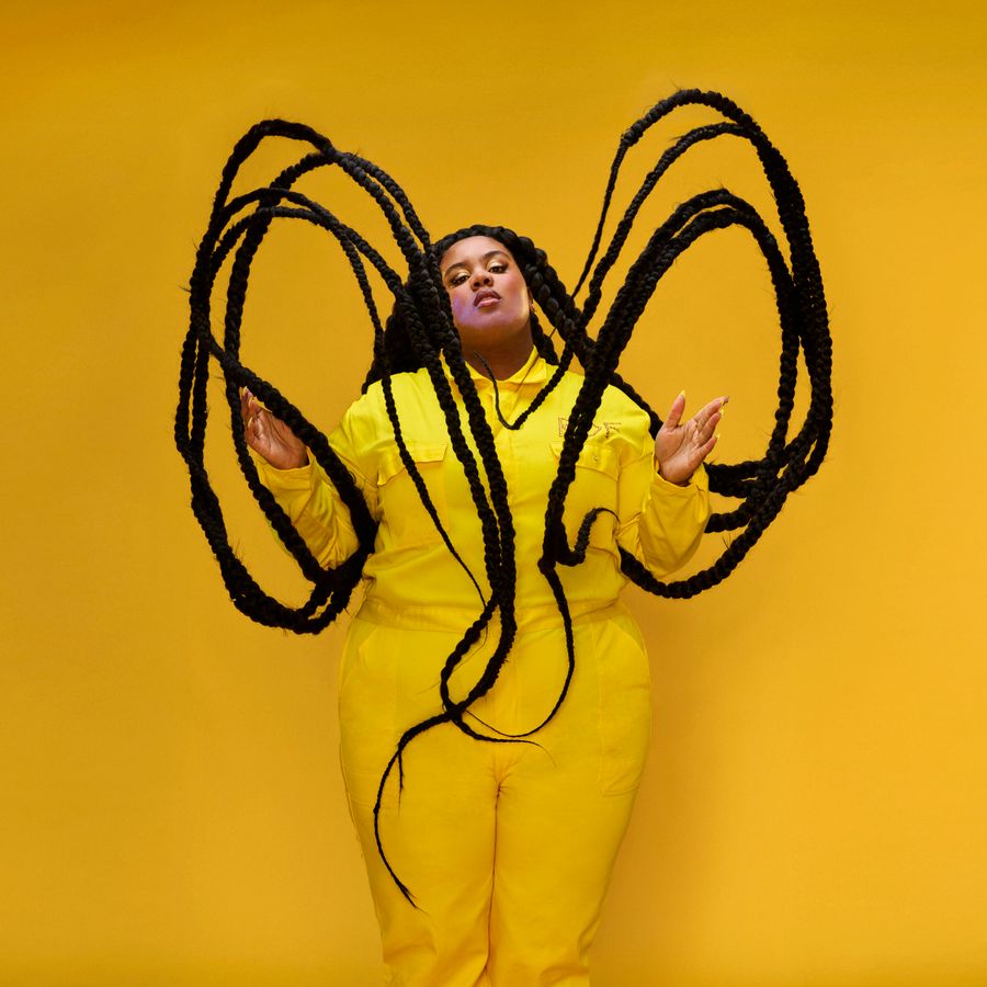 Isabelle Kafando (ook bekend als Rof Daaimund) in een gele overall met lange zwarte vlechten zwevend om haar gezicht