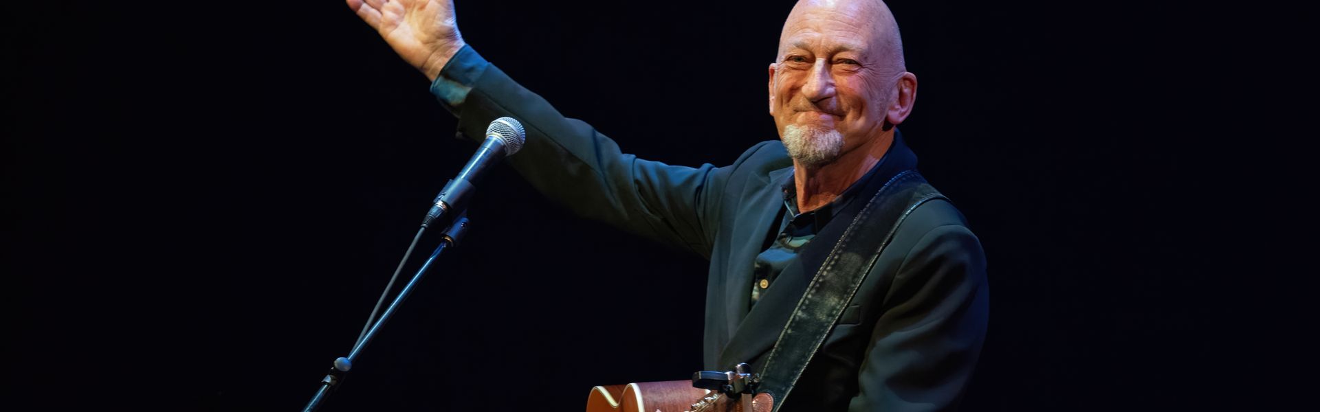 Gerard van Maasakkers met een gitaar achter een microfoon. Hij steekt zijn rechterhand omhoog