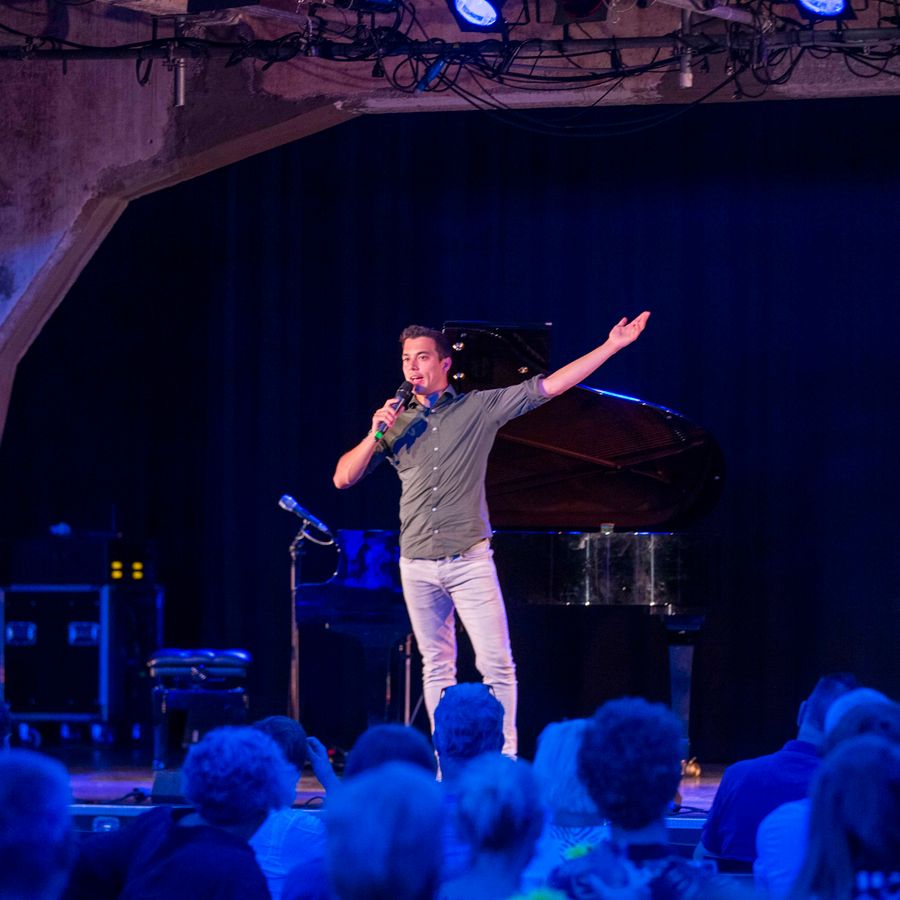Andries Tunru staat op een podium. In zijn rechterhand heeft hij de microfoon voor zijn mond, zijn linkerarm heeft hij wijd gespreid.