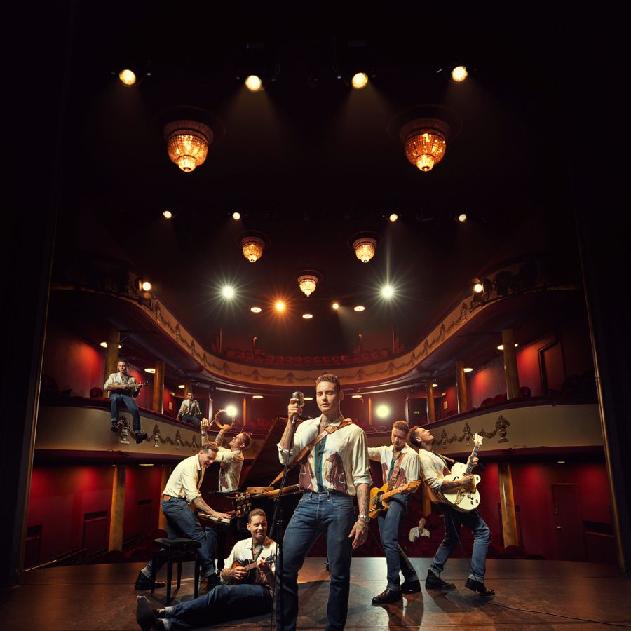 Douwe Bob staat op het podium voor een lege theaterzaal. Om hem heen staan meerdere versies van hemzelf, elk een ander instrument bespelend.
